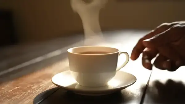 A hand covering a steaming cup of tea with a small white saucer on a wooden table