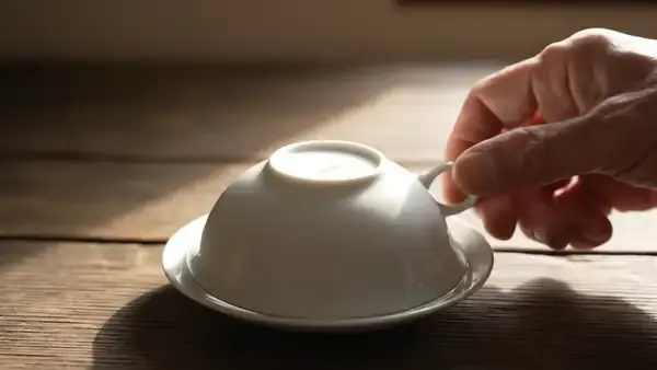 A saucer lifted off a cup to show pale golden tea with unfurled leaves inside